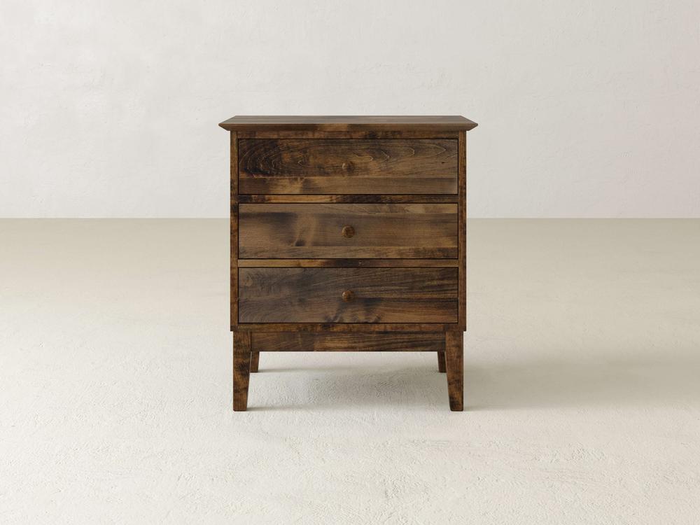 The Bennett 3 Drawer Nightstand by James & James, crafted from solid maple with round knobs, stands on a light floor against a plain, light background. It features a natural wood grain finish and embodies a simple yet sturdy traditional design.