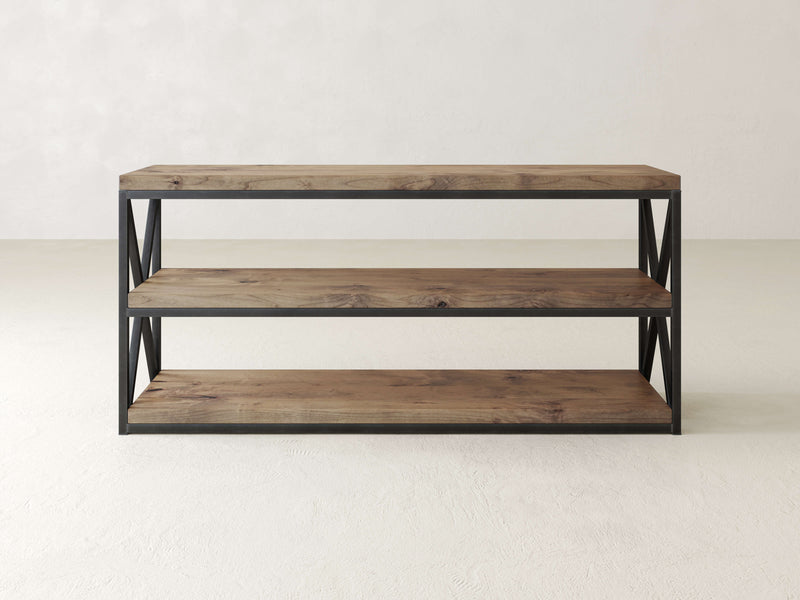 A three-tier wooden shelf with black metal frame and open sides, featuring X-shaped metal details. The shelves are empty, and the unit stands on a light gray floor against a light gray wall.