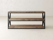 A three-tier wooden shelf with black metal frame and open sides, featuring X-shaped metal details. The shelves are empty, and the unit stands on a light gray floor against a light gray wall.