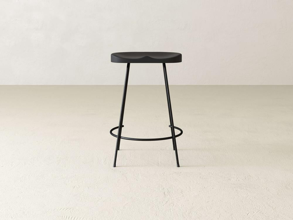 The Bryer Stool by James & James showcases an industrial style with its sleek black round seat and three slender legs on a plain beige floor. A footrest ring links the legs, adding to its modern look against the smooth, light-colored wall.