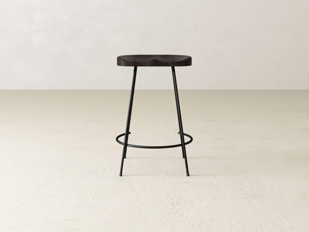 The Bryer Stool by James & James exudes industrial flair with its dark wooden seat and slender black metal legs on a light beige floor. The legs, featuring a circular footrest, enhance its rustic charm against the plain off-white wall.