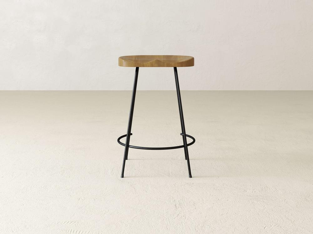 The Bryer Stool by James & James is a minimalist piece with a round wooden seat that exudes rustic charm. It's complemented by slender black metal legs and a footrest, standing elegantly against a plain, light-colored background.