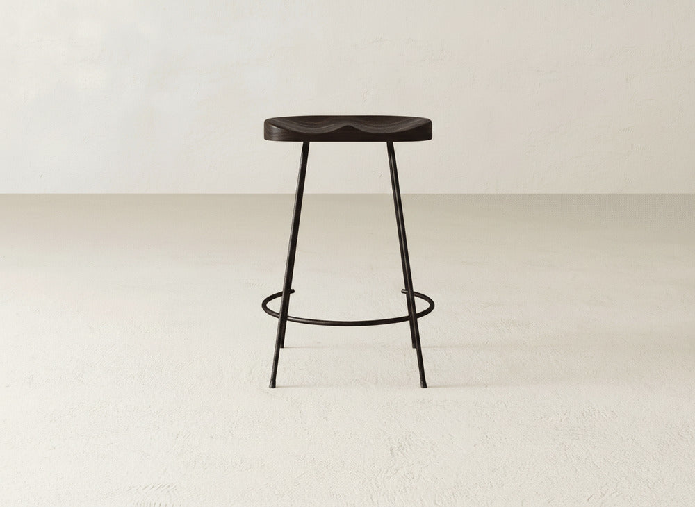 The Bryer Stool by James & James, featuring a dark wooden seat, slim black metal legs, and a circular footrest, stands on a light floor against an off-white wall.