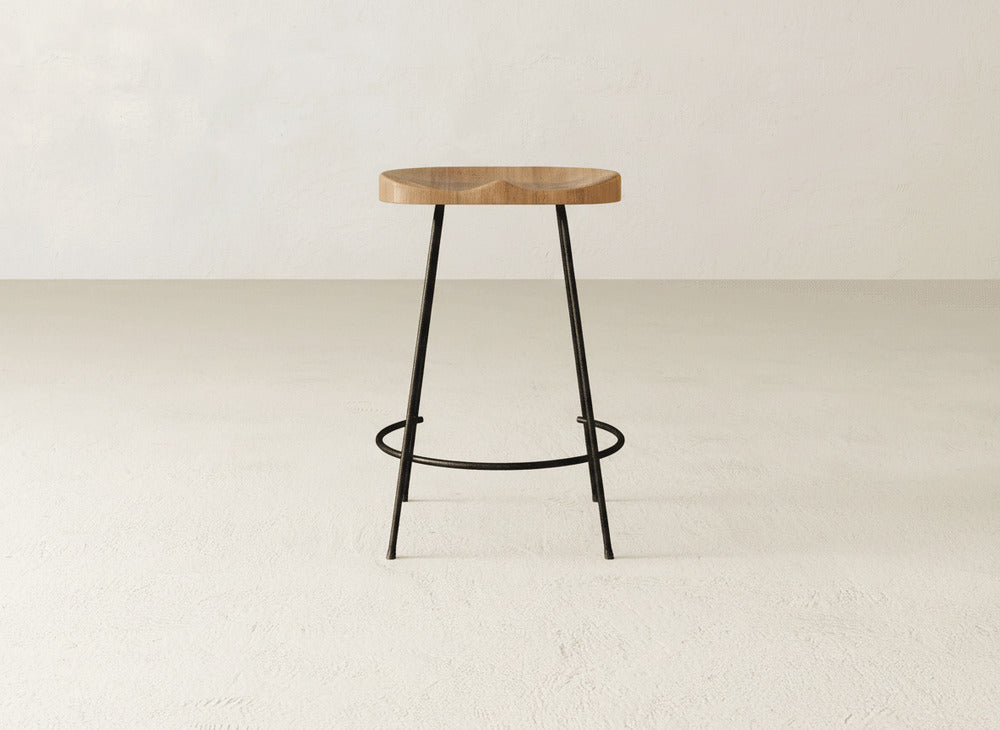 The Bryer Stool by James & James features a minimalist industrial design with a round wooden seat and black metal legs, shown against a plain, light-colored background.
