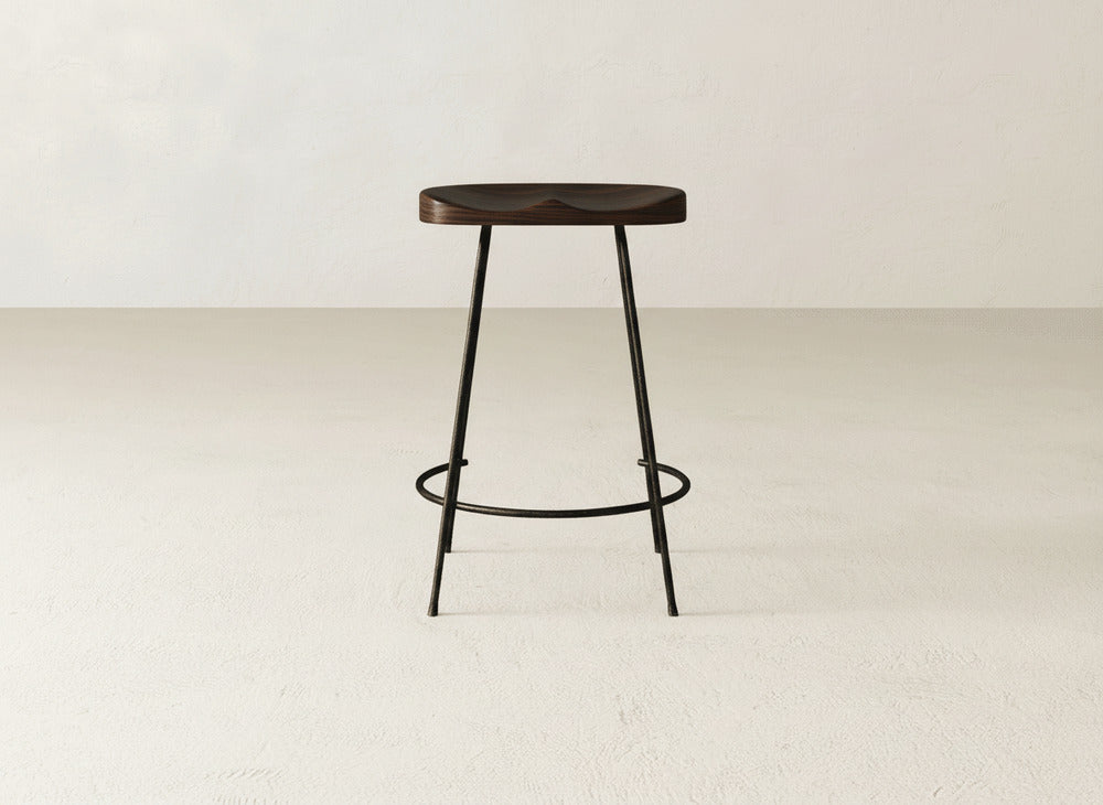 The Bryer Stool by James & James features a dark wooden seat and slim black metal legs, standing alone on a light textured floor against a plain off-white wall for a minimalist rustic look.