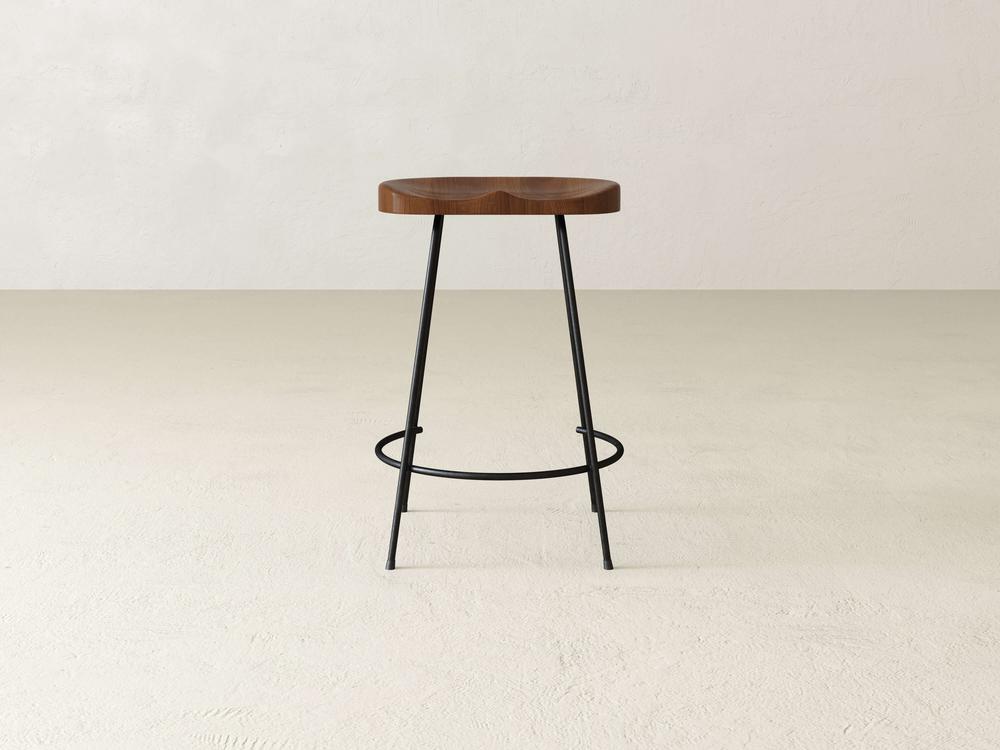 The Bryer Stool by James & James exudes rustic charm with a solid round wooden seat and three slender black metal legs. It stands on a light-colored floor against a textured, off-white wall, featuring a minimalist design accented by an industrial footrest ring connecting the legs.