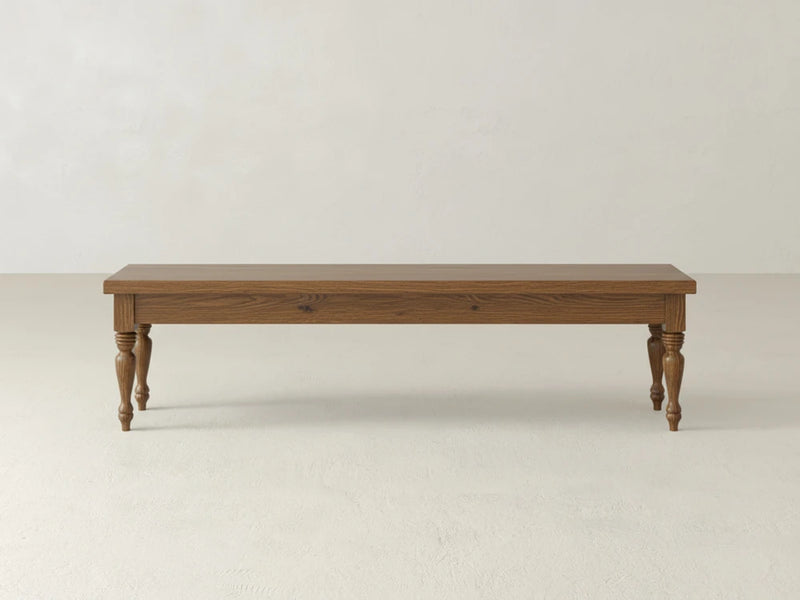 The James & James Chateau Bench - Almond on Oak features a rectangular wooden seat and four turned legs, shown against a plain, light-colored wall atop a pale floor.