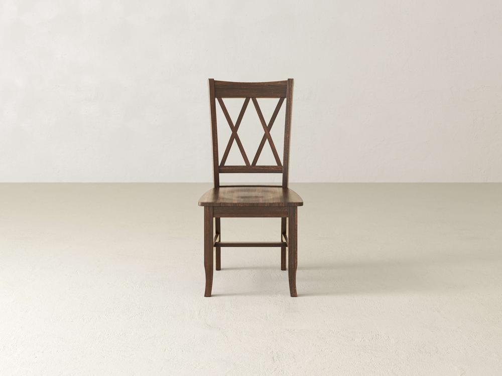 A James+James Double X-Back Dining Chair - Tobacco is placed against a plain, light-colored wall. The chair, featuring an X-shaped backrest design and straight legs, complements the minimalistic setting with an empty, smooth floor.