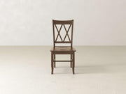 A James+James Double X-Back Dining Chair - Tobacco is placed against a plain, light-colored wall. The chair, featuring an X-shaped backrest design and straight legs, complements the minimalistic setting with an empty, smooth floor.