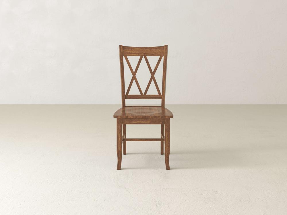 A single Double X-Back Dining Chair - Tuscany from James+James with a decorative backrest featuring an "X" design. The chair is positioned against a light beige wall on a beige floor. Its design is simple and classic, with a warm brown finish.