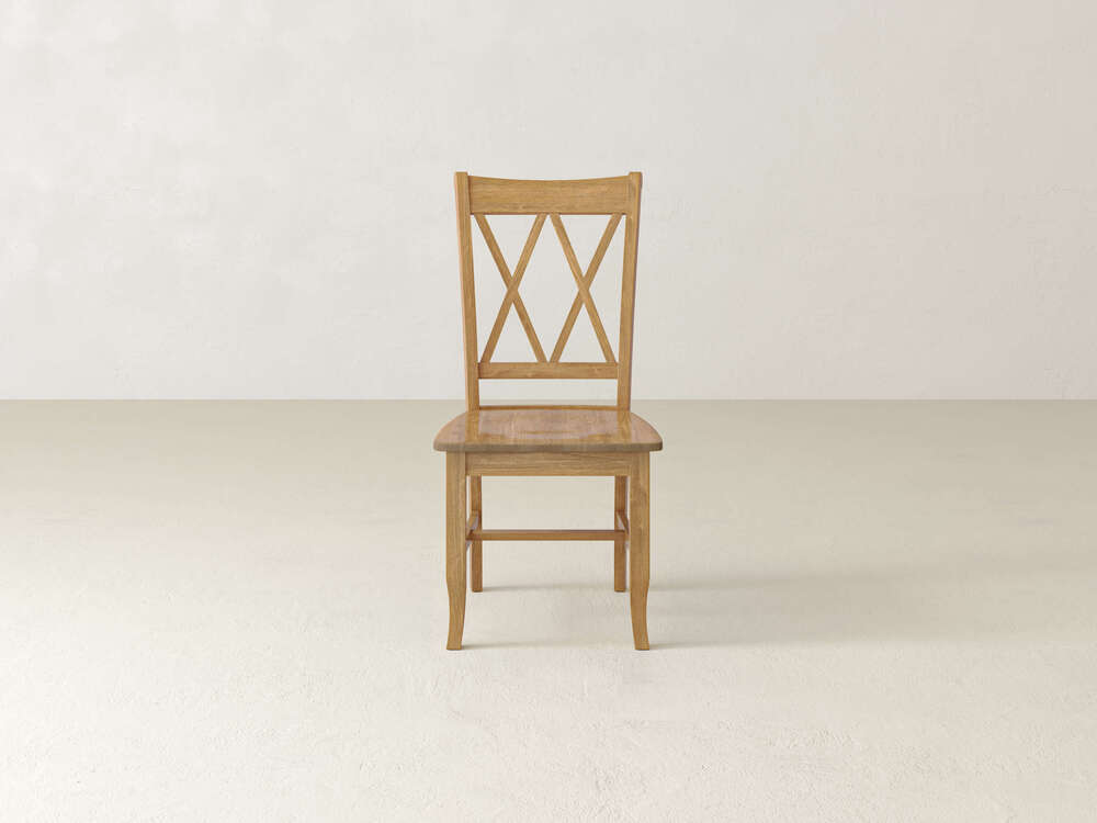 A single Double X-Back Dining Chair - Tuscany from James+James with a decorative backrest featuring an "X" design. The chair is positioned against a light beige wall on a beige floor. Its design is simple and classic, with a warm brown finish.