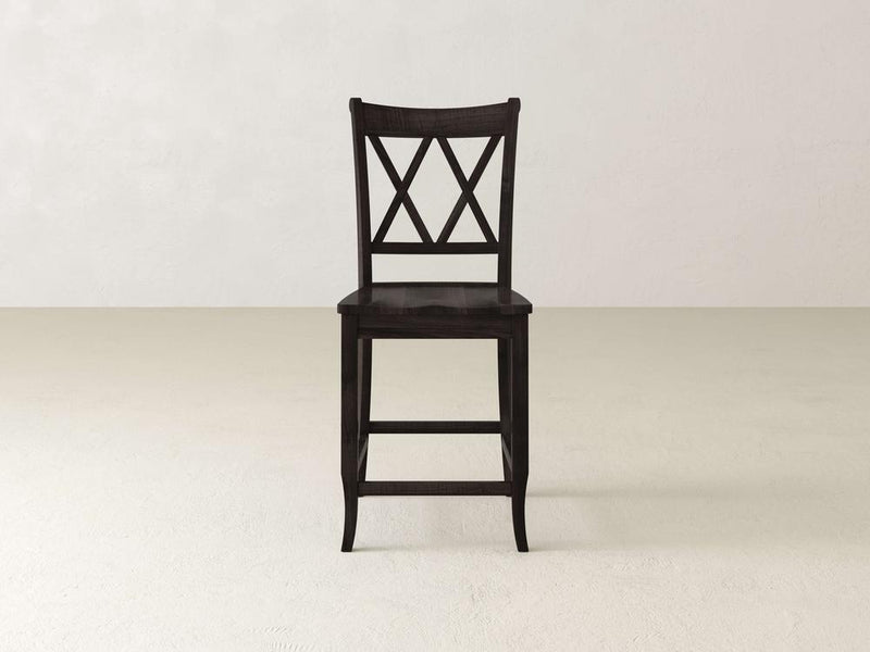 A James & James Double X Back Stool, featuring a dark wood finish, stands on a light floor against a plain white wall. This chair showcases superior craftsmanship by blending classic design with timeless elegance, highlighting its sturdy and refined nature.
