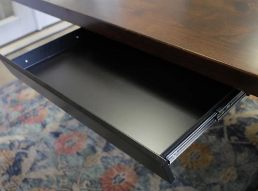 A Standard Desk Add-On by James+James featuring a wooden top with an open black metal drawer beneath it. The drawer is empty, and the desk stands on a colorful, patterned rug with floral designs. In the background, part of a white door and a light-colored floor are visible.