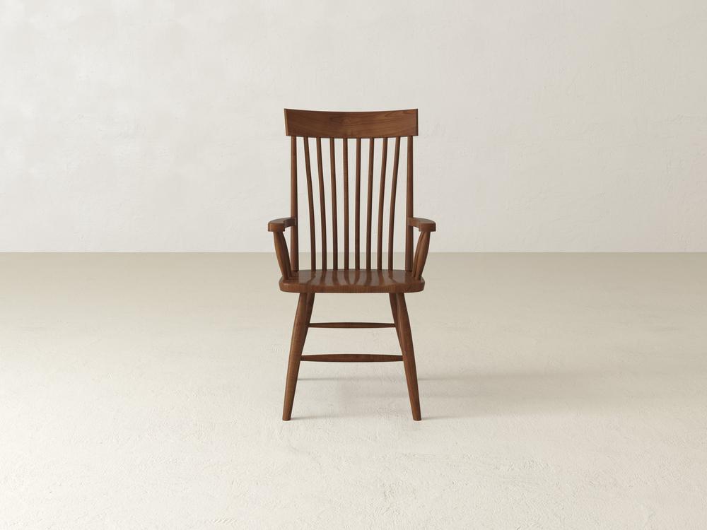 A wooden chair with a high backrest, featuring vertical slats and armrests, is placed on a light-colored floor against a plain white wall. The chair has a minimalist and classic design.