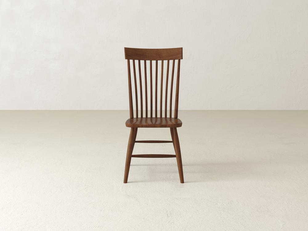 A single wooden chair with a flat seat and a slatted backrest stands on a light-colored floor against a plain off-white wall. The chair has a simple, elegant design with vertical slats and tapered legs.