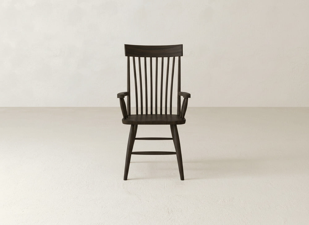 The James+James Eleanor Arm Dining Chair, a dark solid wood piece with a tall vertical slat back and armrests, sits centered in a minimal, light-toned room with plain walls and flooring.