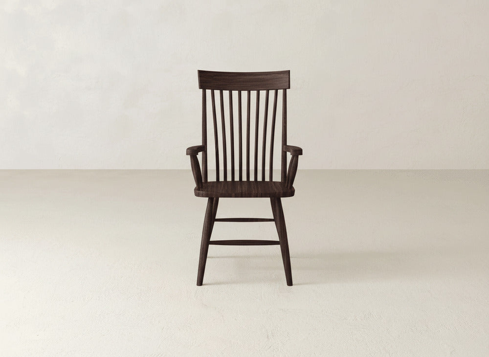 The James+James Eleanor Arm Dining Chair, crafted from solid wood with a high vertical slatted backrest and armrests, stands alone in a minimalist, light-toned room with a plain wall and floor.