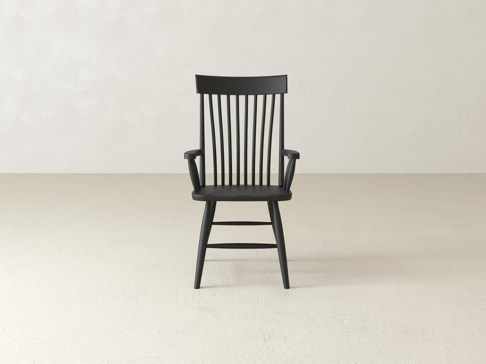 A wooden chair with a high backrest featuring vertical slats, armrests, and a simple design is centered against a plain, light-colored background.