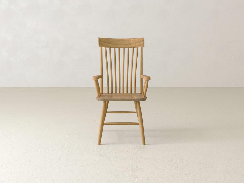 A wooden chair with a high backrest, featuring vertical slats and armrests, is placed on a light-colored floor against a plain white wall. The chair has a minimalist and classic design.