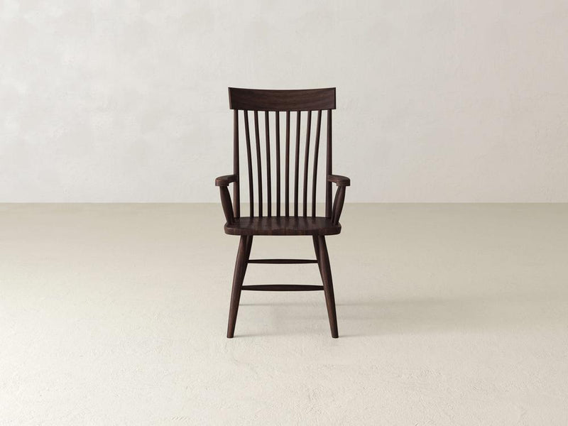 A dark wooden chair with vertical slats on the backrest and armrests is centered in front of a plain, light-colored wall on a smooth, light floor. The chair has a classic, minimalist design.