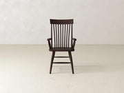 A dark wooden chair with vertical slats on the backrest and armrests is centered in front of a plain, light-colored wall on a smooth, light floor. The chair has a classic, minimalist design.