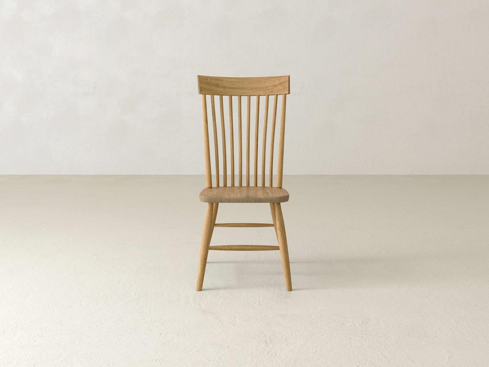 A single wooden chair with a flat seat and a slatted backrest stands on a light-colored floor against a plain off-white wall. The chair has a simple, elegant design with vertical slats and tapered legs.