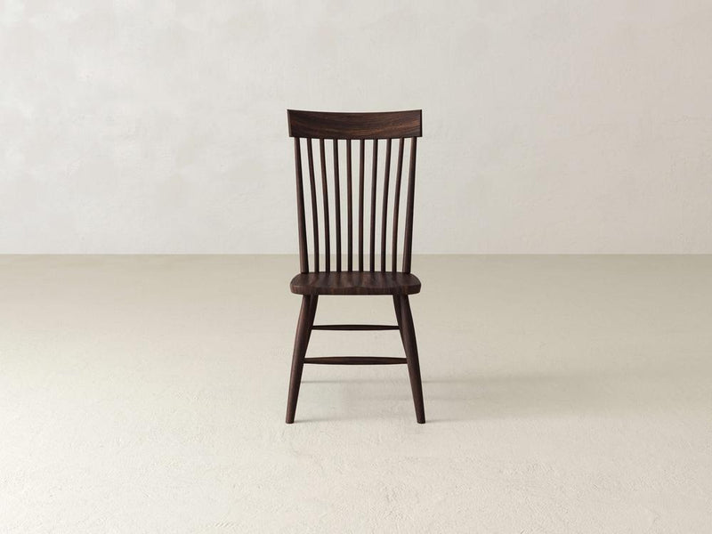 A wooden chair with a dark finish is centered against a plain, light-colored wall. It has a high slatted back and four legs, offering a simple, classic design. The floor is also light-colored, complementing the minimalistic setting.