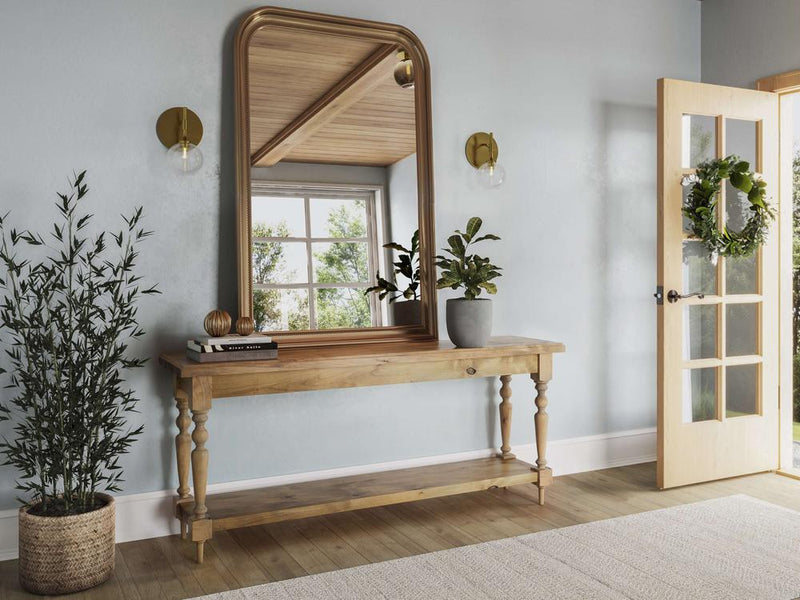 A bright entryway features the Ella Sofa Table in Harvest Wheat by James+James, adorned with a potted plant, books, and decorative objects. Above the table hangs a large arched mirror. A plant in a wicker basket sits to the side. An open door with a wreath invites sunlight.