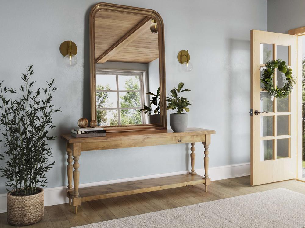 A bright entryway features the Ella Sofa Table in Harvest Wheat by James+James, adorned with a potted plant, books, and decorative objects. Above the table hangs a large arched mirror. A plant in a wicker basket sits to the side. An open door with a wreath invites sunlight.