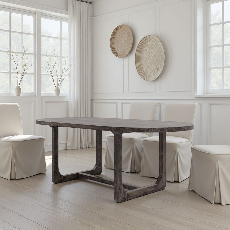 A modern dining room features the James+James Emersen Oval Dining Table in Tobacco, four white slipcovered chairs, large windows with sheer curtains, woven baskets on a white wall, light wood floors, and minimal decor for an airy vibe.
