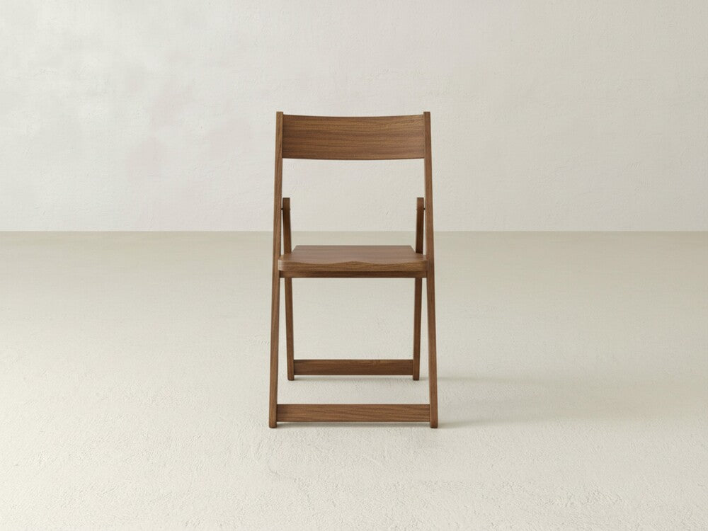The Emmett Folding Chair by James & James, featuring a straight backrest and solid maple hardwood seat, is set on a light floor against an off-white wall.
