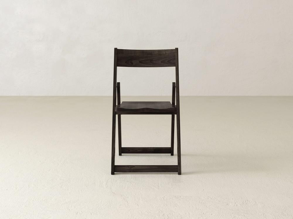 A minimalist black folding chair with a gray fabric seat is placed on a plain, light-colored floor against a bare, off-white wall. The chair is centered and viewed from the front.