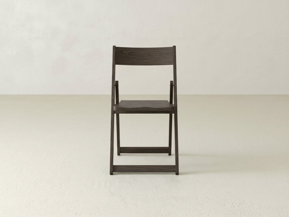 The Emmett Folding Chair by James & James, made from solid maple hardwood with a dark finish and wooden seat, is centered in an empty room with light walls and a textured floor, viewed from the front.