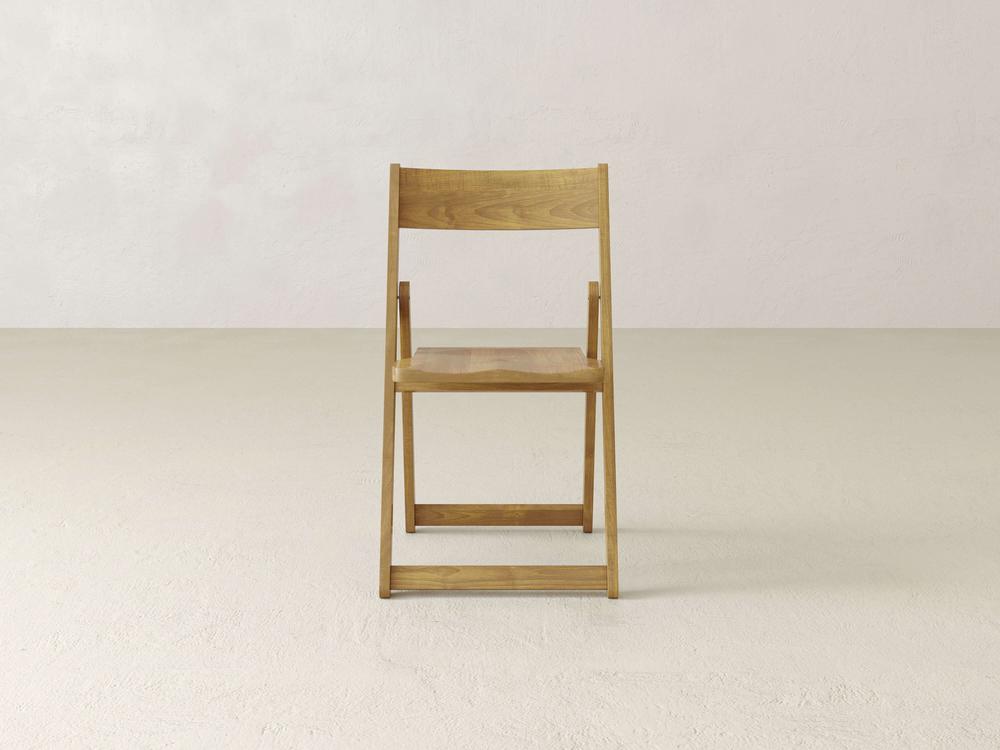 A single dark wooden folding chair sits on a light-colored floor against an off-white wall. The chair is positioned facing forward and centered in the minimalist setting.