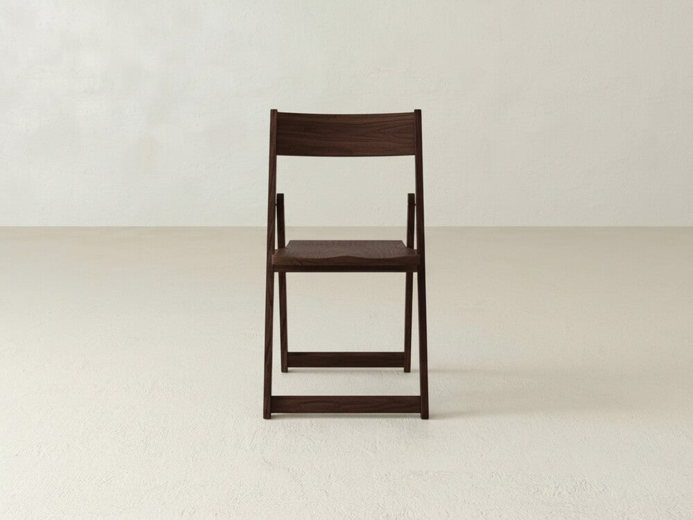 The James & James Emmett Folding Chair, with its dark wood and wooden seat, is centered in a minimalist room with a light floor and plain white wall backdrop.