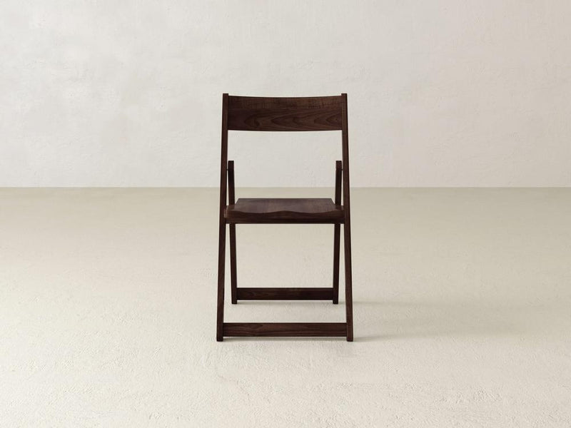 A wooden folding chair with a dark finish is placed on a light beige floor against a plain off-white wall. The chair is centered and viewed from the front.