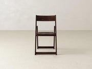 A wooden folding chair with a dark finish is placed on a light beige floor against a plain off-white wall. The chair is centered and viewed from the front.