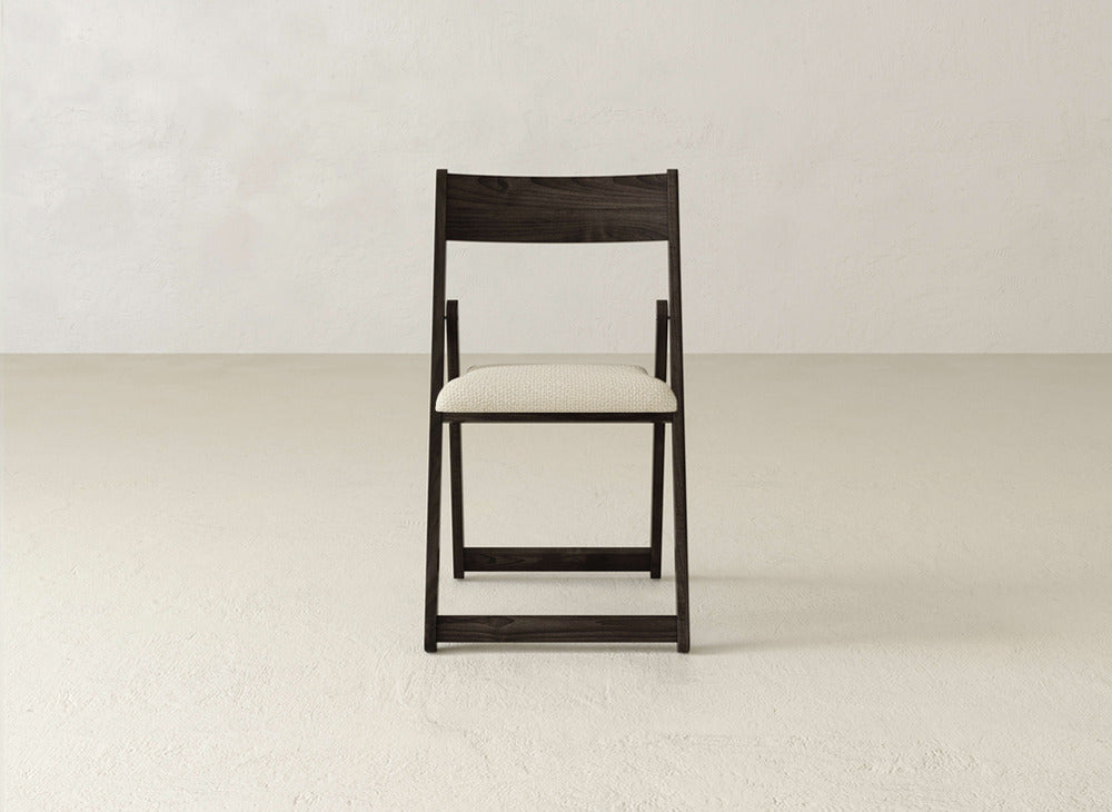The Emmett Upholstered Folding Chair by James & James, featuring a dark wood frame and light seat, is placed in the center of a simple, light-toned room with textured walls and flooring.