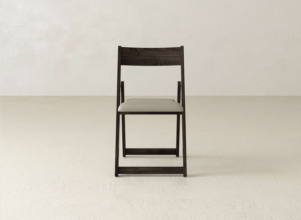 The Emmett Upholstered Folding Chair by James & James, featuring dark wood and a light cushioned seat, is centered in an empty room with light beige walls and a matching floor.