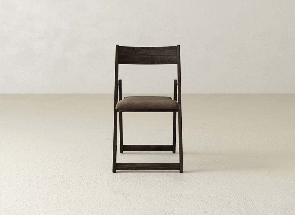 The Emmett Upholstered Folding Chair by James & James, featuring a solid maple hardwood frame and cushioned brown seat, is centered in an empty, light-colored room with textured walls and floor.