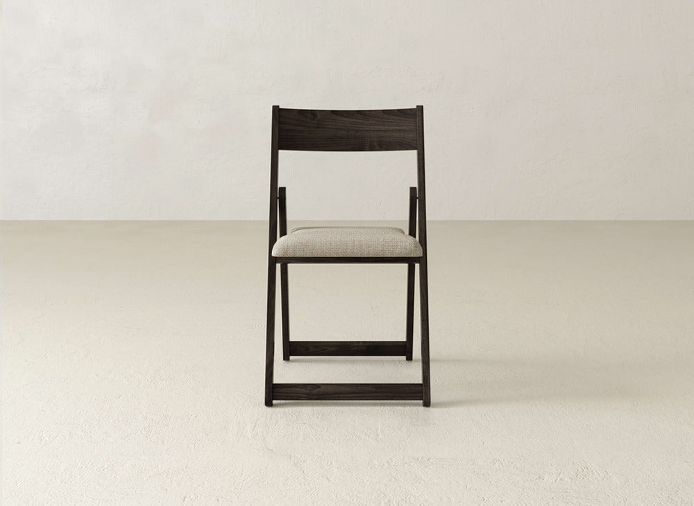 The James & James Emmett Upholstered Folding Chair, featuring a dark solid maple frame and light beige cushioned seat, is placed at the center of a minimalist room with plain, light-colored floors and walls.