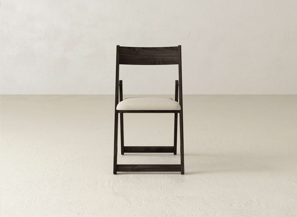 The Emmett Upholstered Folding Chair by James & James, featuring a dark wood frame and light beige cushioned seat, is centered against a plain, light-colored wall and floor.