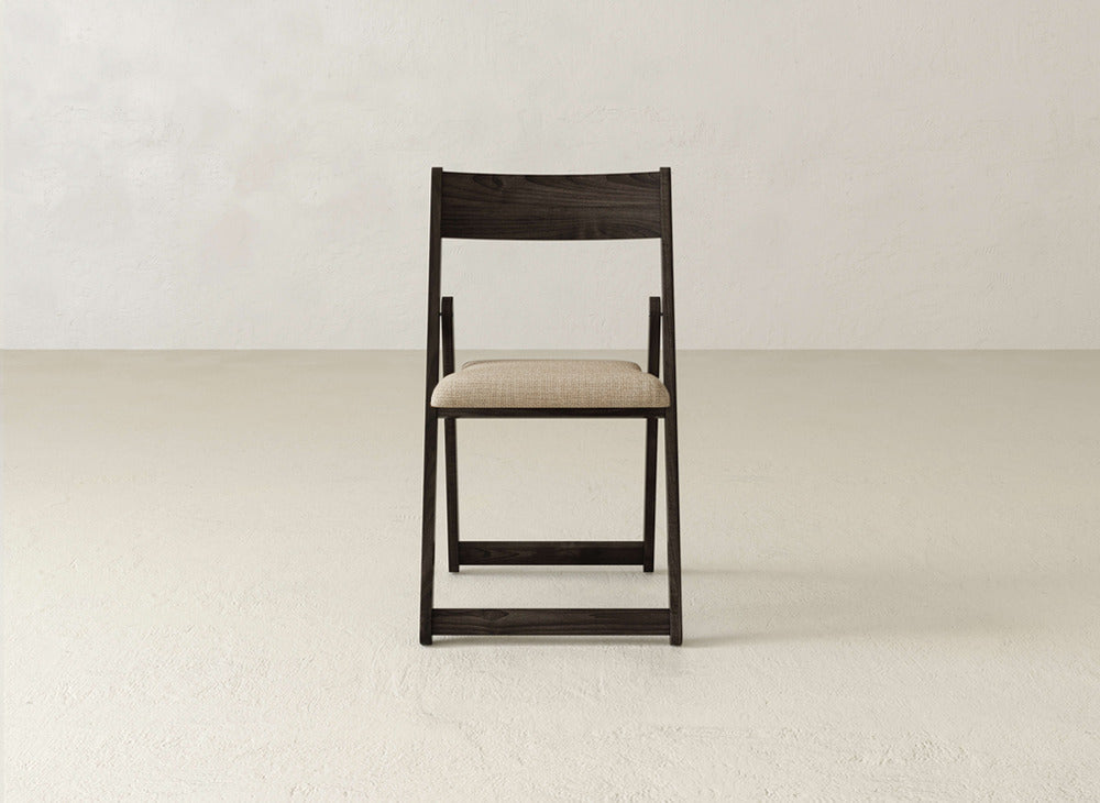 The Emmett Upholstered Folding Chair by James & James—a dark solid maple chair with a light beige cushioned seat—sits centered on a plain, light-colored floor against a neutral wall, facing forward. Its practical foldable design offers versatility and style.