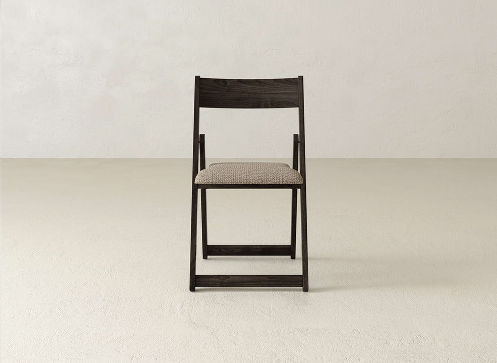 The Emmett Upholstered Folding Chair by James & James features a dark wood frame and beige cushioned seat, centered in an empty room with light walls and floors to highlight its practical, foldable design.
