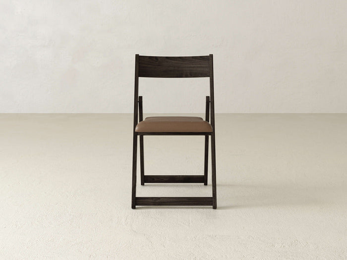 The Emmett Upholstered Folding Chair by James & James, featuring a brown padded seat, sits centered and facing forward on a light textured floor against a plain off-white wall.