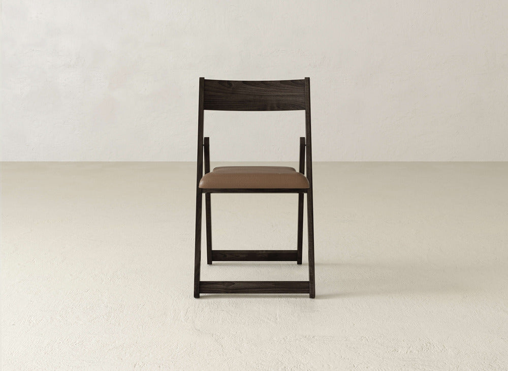 The Emmett Upholstered Folding Chair by James & James, featuring a brown padded seat, sits centered and facing forward on a light textured floor against a plain off-white wall.