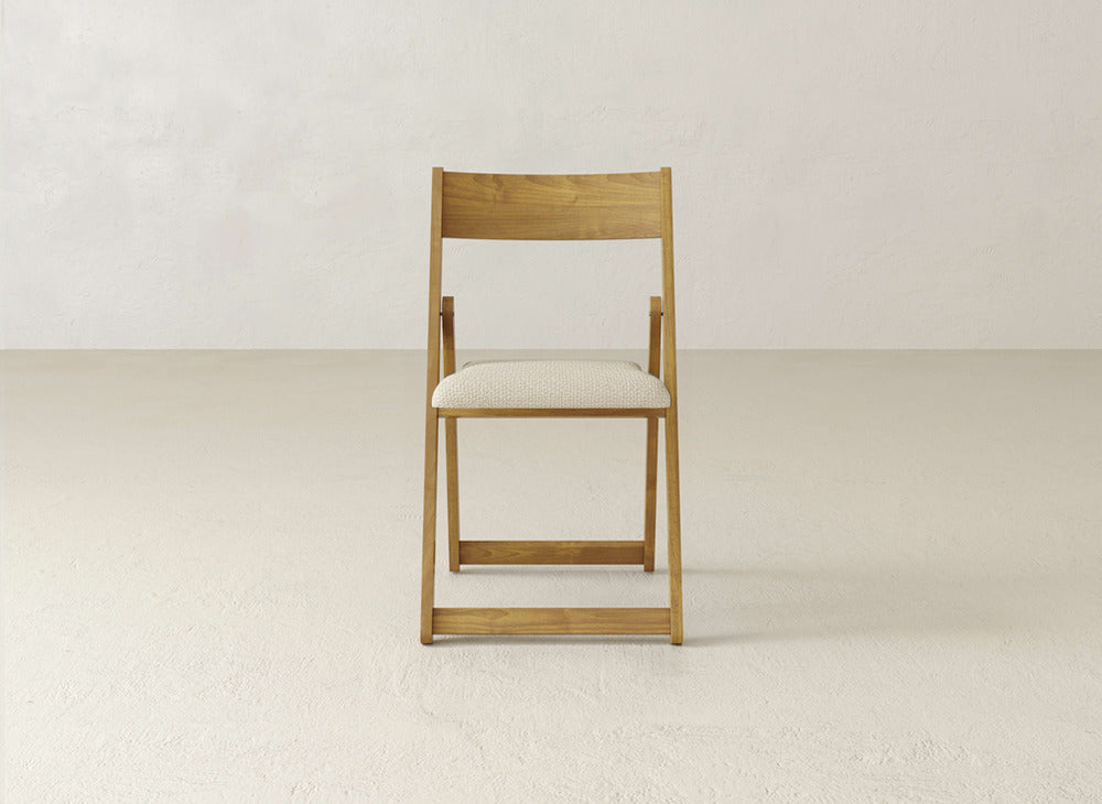 The Emmett Upholstered Folding Chair by James & James, featuring maple hardwood and a light cushioned seat, stands centered against an off-white wall and floor, highlighting its elegant design.