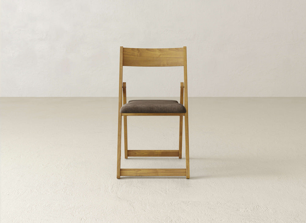 The Emmett Upholstered Folding Chair by James & James, featuring a maple hardwood frame and brown cushioned seat, is positioned front-facing on a light floor against a beige wall.