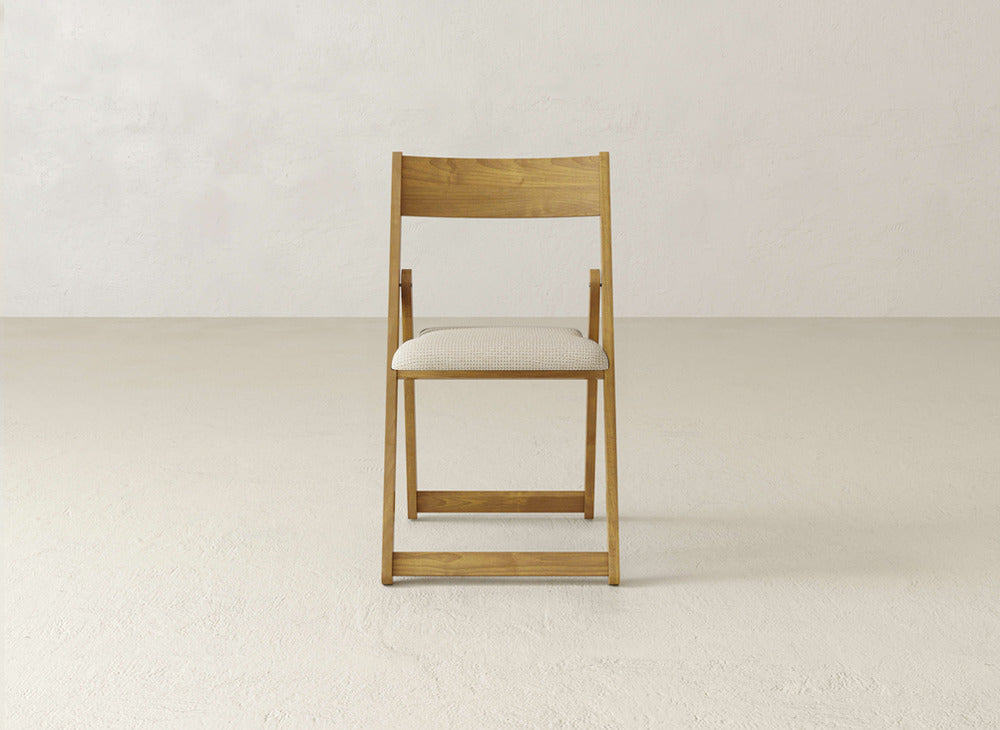 The Emmett Upholstered Folding Chair by James & James, featuring a light beige cushioned seat and a solid maple hardwood frame, sits in the center of an empty room with plain, light-colored walls and floors.
