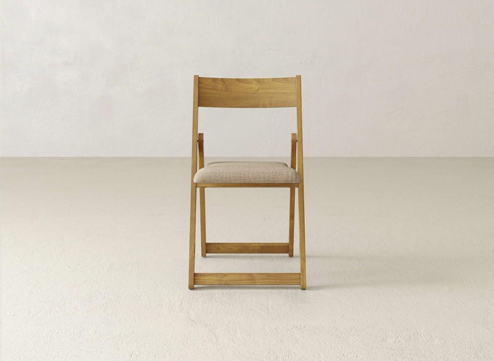 The Emmett Upholstered Folding Chair by James & James, featuring a padded beige seat, sits on a light floor against a plain white wall.